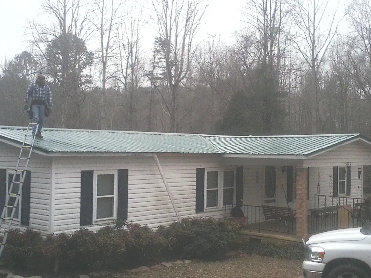 Worker on a ladder beside a new green metal roof on a ranch home in Willoughby
