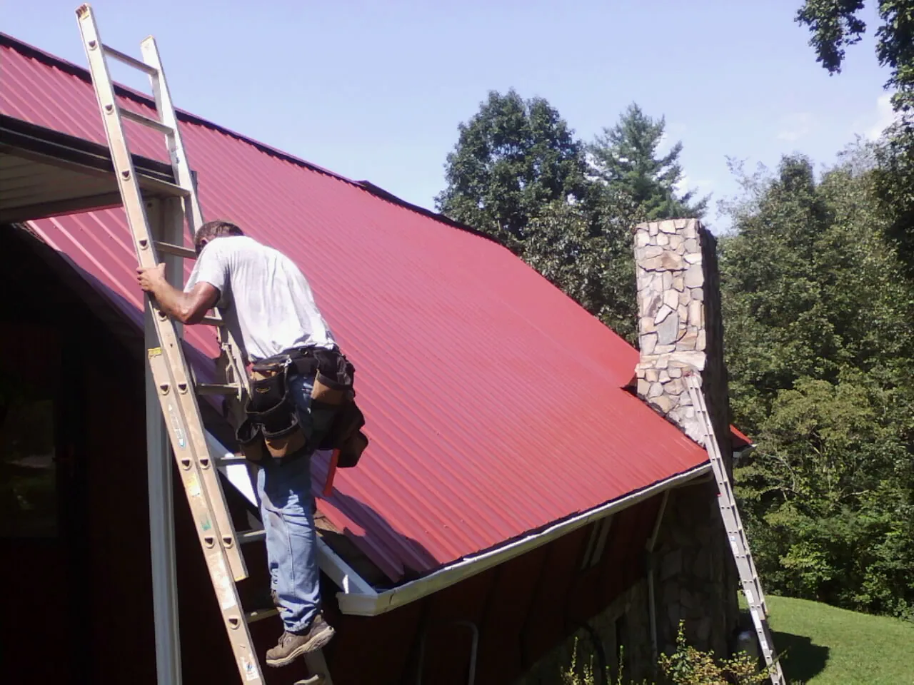Roofer on a ladder beside a red metal roof and stone chimney — Soffit & Fascia Repair by Roofing Brothers in Willoughby